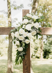 Wooden cross adorned with white roses, baby's breath, and greenery for an outdoor wedding ceremony at Ashford Acres Inn.