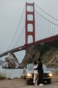 A couple embraces in front of a vintage car during a photoshoot with the Golden Gate Bridge in the background, enveloped in a misty atmosphere in San Francisco.
