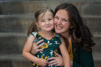 A woman and a young girl smile warmly at the camera, sitting on stone steps. The girl wears a floral dress; the woman, a green cardigan. The mood is joyful.