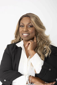 Confident headshot of a woman in a black blazer and white blouse, smiling with poise against a white backdrop—created to showcase leadership and personal branding strength.