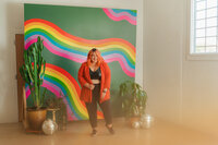A person with vibrant hair smiles standing in front of a rainbow mural