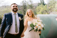 Groom and bride smile while holding hands and with a flower bouquet outside