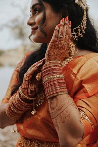 A close-up of one of the marriers at an Indian wedding in Phoenix, Arizona. They are dressed in an orange sari adorned with gold jewelry, showing off intricate henna designs on their hands, with a soft smile on their face.