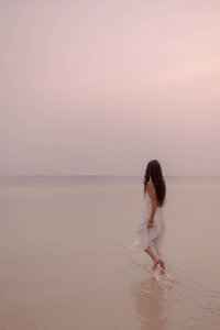 Courtney walks away from the viewer in a white dress in the ocean, dreamily gazing off into the hazy pink horizon, representing creative storytelling