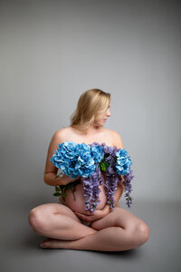 Blonde pregnant woman showing maternity belly while sitting in neutral studio and holding blue and purple flowers.  