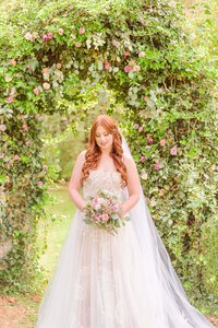 A bride smiles softly as the light streams through the leafy arch behind her.