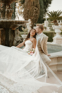 Photo taken by Bailee Freeman Photography of a bride and groom sitting on the edge of a fountain in front of the Grand Island Mansion in Walnut Grove, California. Bride is in front of the groom and leaning in while the groom kisses her forehead. 
