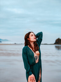 Woman with long hair stands at the edge of the ocean in a dark green cardigan – dreamy adventure branding portrait by Mewphotovideo on the Oregon Coast