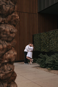 Romantic couple embracing outdoors against a sleek modern building exterior with green hedges and warm brown walls in downtown Phoenix, Arizona, captured in a stylish urban engagement photoshoot by a local Phoenix wedding photographer