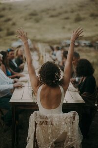 A bride sits at the head of a long narrow table with her back to the camera and her hands raised. Several people are sitting at the table looking at each other in conversation.