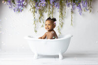 A joyful toddler sitting in a white vintage-style clawfoot tub surrounded by delicate bubbles, smiling brightly against a whimsical floral backdrop of purple wisteria and greenery. The pristine white setting and vibrant child's expression create a dreamy, playful scene perfect for a milestone photoshoot. Captured by a talented Dallas milestone photographer.