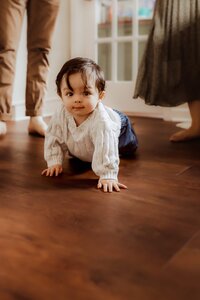 A baby in a white sweater and jeans crawls on a wooden floor, while three adults stand nearby.