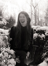 A 120 film black-and-white photograph from a branding session for a fine foods store. The image captures a candid moment of a woman sitting outdoors, laughing warmly, surrounded by snow-covered bushes and floral arrangements. The natural light enhances the timeless, organic feel of the scene, perfectly reflecting the store’s authentic and inviting brand identity.