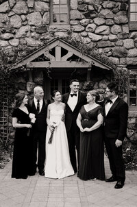 Black and white film photo of a couple with their family in front of the stone facade at Willowdale Estate during a winter wedding — a joyful portrait made by a film wedding photographer.