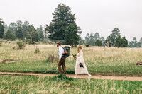 couple walking on a path with a back pack, hats and a bouquet. There is tall grass and green pine trees around them 