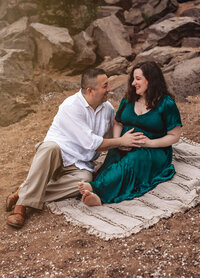 Toddler stands at the end of a long twisting lane that curves behind her. There's snow on the ground and tall brown grass behind her with trees in the background. She is wearing a tan coat and white snow hat with a pompom and is smiling big at the camera in this family photoshoot.