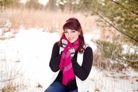 A red headed high school senior holding her bright pink scarf around her neck while kneeled down in the snow