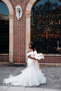 the bride stands in the courtyard of the wedding venue, holding her flowers and looking down at her gown's train