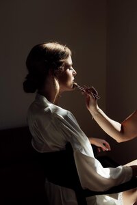 Bruidsfotografie: moment tijdens de voorbereidingen waarin bruid haar make-up krijgt aangebracht in sfeervol zijlicht