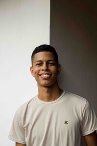 A young Black teen with short hair, wearing a beige t-shirt, smiles toward the camera. They are leaning against the corner of two intersecting white walls.