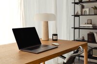 A wooden desk with an open laptop, mouse, and a beige lamp with a matching shade. A candle sits in the back right corner, and white net curtains hang in the background, diffusing natural light.
