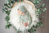 Newborn baby swaddled in a sage green wrap with a matching bonnet, resting on a flokati rug atop a wooden prop, surrounded by soft eucalyptus leaves.