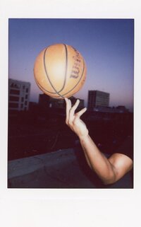 Instax instant film by Renata Voci showing Jordan Foster's hand balancing a basketball on the tips of his fingers on a rooftop in LA. Harsh flash shows the ball brightly while the background is dark blue hour lighting.