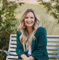 Cheerful woman with long blonde hair, wearing a teal blazer and coral earrings, sitting on a silver bench in a grassy outdoor area with trees and red berries in the background.