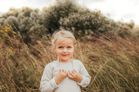 little girl tugs her pigtails while the wind blows and softly smiles, in a grassy field.