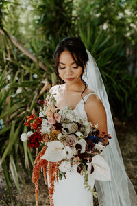 Bride standing in front of lush green trees, holding a wedding bouquet with lilies