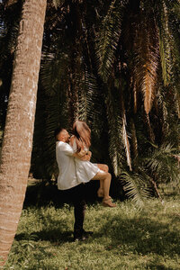 Man lifting his partner with laughter beneath towering palm trees, captured during a playful and romantic couples session in a tropical Florida setting.