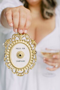 Woman in a white dress holding a champagne coupe and a gold emblem that reads "press for champagne" above a doorbell
