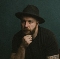 Man with a red beard and tattoos, dressed in black and wearing a wide-brimmed hat, seated against a moody green backdrop with a pensive expression.