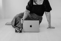 black and white photo of photographer Bethany Simms with her laptop and a camera sitting on the floor