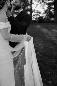 A black and White photo of a Bride wearing lace-beaded bridal gloves holding up her beaded veil as it drapes to the ground.