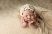Newborn baby girl posed with her head in her hands wearing a floral bonnet .