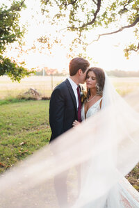 A romantic scene of a bride and groom at sunset, their figures outlined by the warm hues of the evening sky, veil shot.