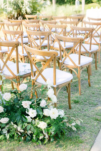 Wooden cross-back chairs with white cushions flank lush white floral aisle arrangements at an outdoor Bluffton wedding ceremony—captured by luxury wedding photographer Amia Marcell.