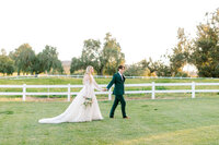 Bride and groom walk hand in hand on a grassy field surrounded by white fences and distant trees under a clear sky.