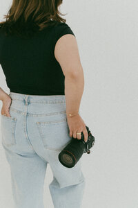 Rear view of a woman in jeans and a black top holding a camera lens in her right hand with a clean studio backdrop.