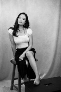 model posing casually for a portrait. She is sitting on a stool, wearing a long black skirt and a beige top. The image is in black and white.