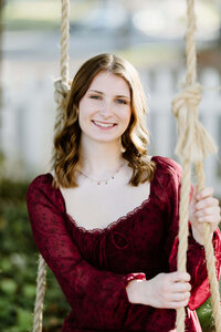 senior portrait of 2023 Hallsville graduate in maroon dress swinging on porch swing at bed and breakfast in Jefferson TX