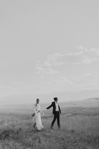 bride and groom holding hands while walking through a field