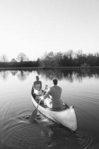A couple paddles a canoe on a tranquil lake, surrounded by trees, captured in a serene black-and-white setting.