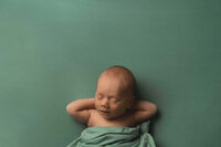 A newborn photograph by Mobile, Al Newborn Photographer of a baby boy sleeping peacefully on a sage green backdrop with his hands tucked comfortably behind his hand with a matching sage green swaddles draped over his body for warmth.