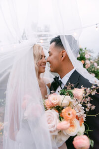 Bride and groom sharing a joyful moment under a veil with blush and peach florals, captured by Austin-based editorial wedding photographer Erin Marcin.
