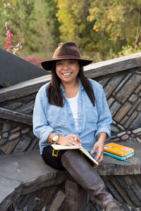 A woman in a denim shirt and brown hat sits outdoors on a stone bench, smiling with a journal in hand. A relaxed, natural setting for professional headshots.