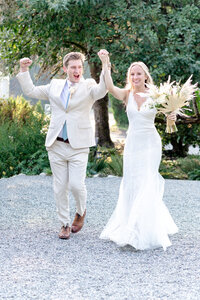 This vibrant, joyful photograph captures a newly married couple celebrating as they walk hand-in-hand down a gravel path. The groom, dressed in a light beige suit, has his fist raised high in excitement, his expression radiating happiness and energy. The bride, in a stunning lace gown, carries a bouquet of dried florals and pampas grass, beaming as she walks alongside him. The lush greenery in the background adds to the natural, outdoor setting, and the movement of their walk gives the image a sense of freedom and celebration, making it a perfect moment of post-ceremony bliss.