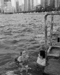 Two people are swimming in a choppy urban waterfront. A worn metal ladder is visible on the right. They appear happy, with a city skyline in the background.