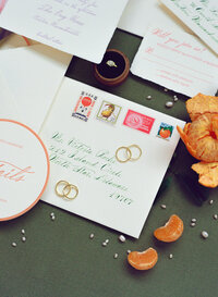 Flatlay of wedding invitation suite on film featuring vintage stamps, calligraphy, gold rings, citrus slices, and scattered pearls — photographed by a film wedding photographer.